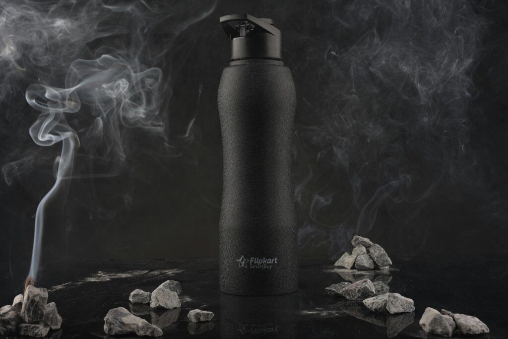 Black plastic bottle surrounded by smoke and rocks in a monochrome setting.